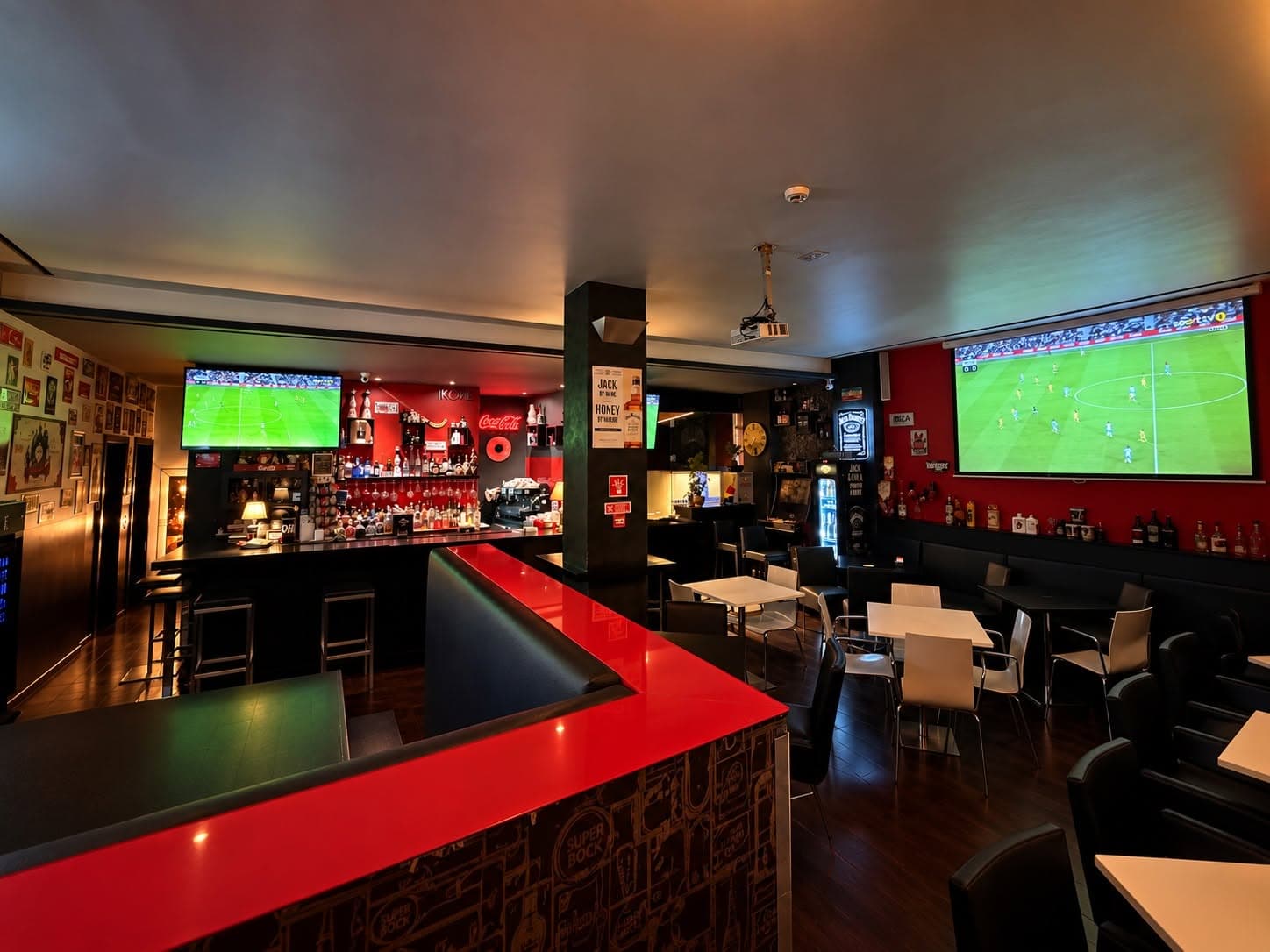 Sala do IKONE bar com balcão vermelho, televisão e projetor a transmitir futebol